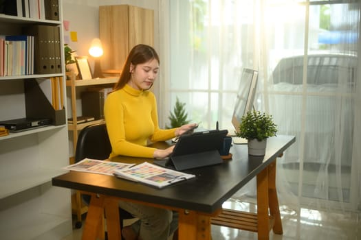 Young female graphic designer working on digital tablet and reviewing color samples at desk.