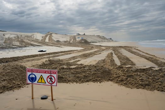 Signs indicate restricted access due to construction efforts aimed at managing coastal erosion while heavy machinery operates on the sand at Biscarosse beach under a cloudy sky.