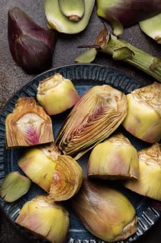 Plate full of raw artichoke hearts on dark rustic table top view. Fresh vegetables bought on farmers market. Cleaning and preparing to cook. Italian cousine ingredient