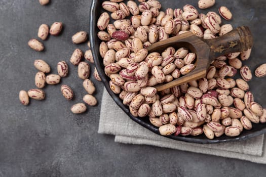 Ceramic plate full of dried pinto beans with a wooden scoop on napkin on gray table top view, copy space. Healthy vegetarian food, legumes protein source. Latin American cousin ingredient