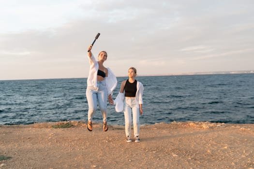 Two women in white shirts and jeans are jumping on a beach, one holding a wooden bat in the air. The ocean and a bright sky are in the background.
