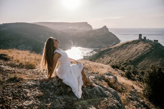 A woman in a white dress is sitting on a rock overlooking a body of water. She is enjoying the view and taking in the scenery