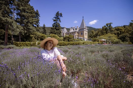 A woman is sitting in a field of purple flowers. The field is surrounded by trees and a large house can be seen in the background. The woman is wearing a straw hat and a white shirt