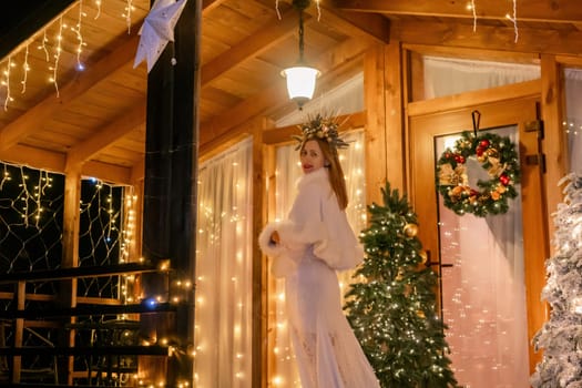 A woman in a white fur coat stands in front of a wooden cabin decorated with Christmas lights and a wreath.