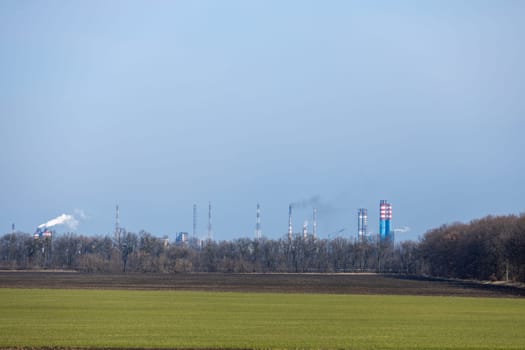 Smoke rising from an industrial plant behind a green field and forest, highlighting environmental impact