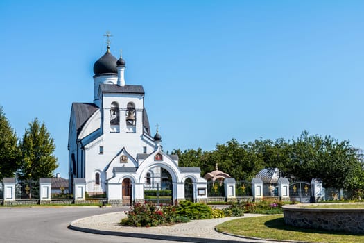 White orthodox church with black domes and golden crosses, standing tall against a clear blue sky, surrounded by green trees and a paved area