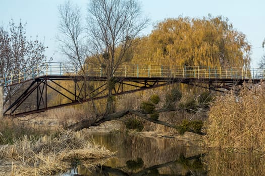 Rusty metal bridge featuring yellow railing, reflecting in calm river water amidst a serene autumn landscape filled with colorful trees