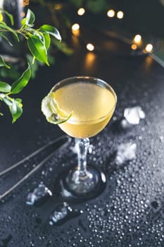 Elegant coupe glass filled with bright yellow cocktail, garnished with a lime twist, sits on a dark, wet bar top, illuminated by warm string lights