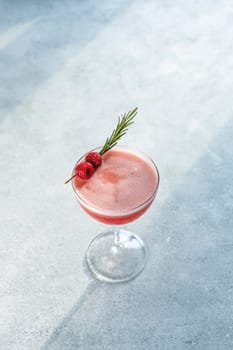 Coupe glass containing a vibrant pink cocktail garnished with fresh raspberries and a sprig of rosemary, creating an elegant and refreshing drink