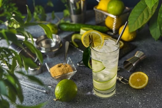 Cucumber lemonade cocktail being prepared by bartender with fresh ingredients, metal straw, and bar tools on grey countertop