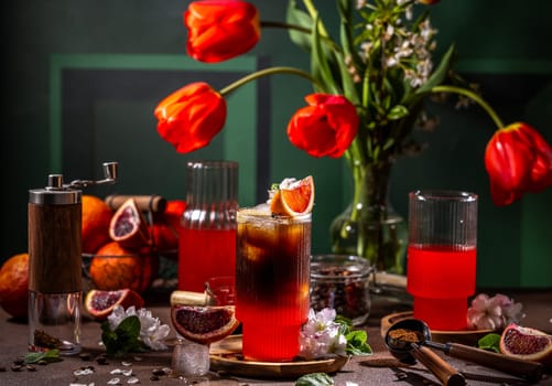Glass of iced coffee with blood orange juice decorated with slice of orange and mint leaves, grinder, red tulips and ingredients on table