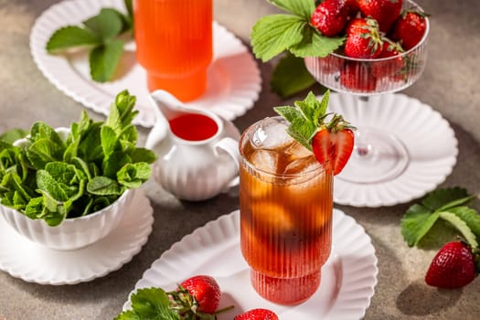 Glass of strawberry iced tea decorated with fresh mint and strawberries, accompanied by ingredients on a table
