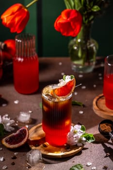 Vibrant red orange and coffee beverage is being served in a stylish glass with ice, garnished with a slice of red orange and delicate white flowers