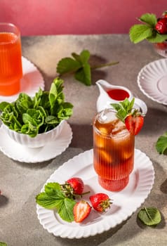 Glass of strawberry iced tea decorated with fresh mint and strawberries, accompanied by ingredients and other beverages on a table