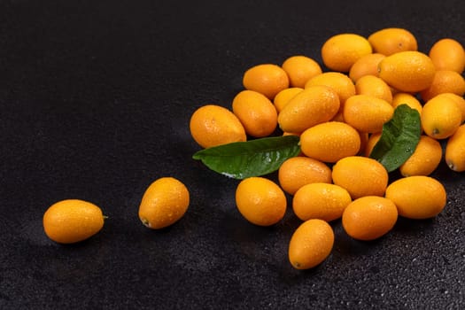 Vibrant kumquats, adorned with water droplets and accompanied by green leaves, create a striking contrast against a dark, wet backdrop