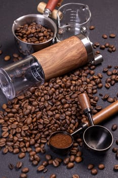 Roasted coffee beans scattered on a dark surface, accompanied by a manual grinder, metal scoop, and a measure of ground coffee, creating an aromatic tableau