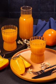Glasses and bottle of persimmon juice served with fresh fruits and decorative flowers on dark background, creating a vibrant and appetizing scene