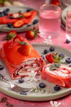 Pink strawberry crepe roll is being sliced and served on a plate with fresh strawberries and blueberries