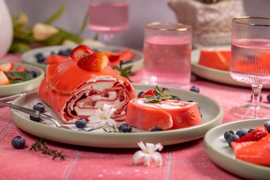 Sliced pink strawberry cake roll filled with cream cheese, adorned with fresh strawberries, blueberries, and white flowers on a pink tablecloth