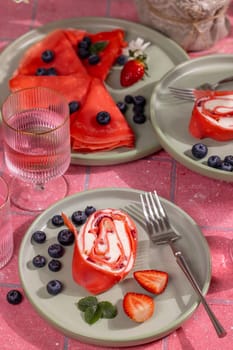 Delicious red pancakes are rolled and filled with cream cheese, decorated with strawberries and blueberries, served on green plates on a pink tiled table