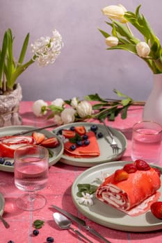Delicious pink strawberry crepes and rolls served on green plates on a pink tiled table decorated with spring flowers and glasses of water