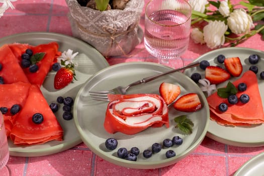 Pink rolled crepe filled with cream cheese and strawberries, paired with red pancakes, fresh berries on vibrant green plates
