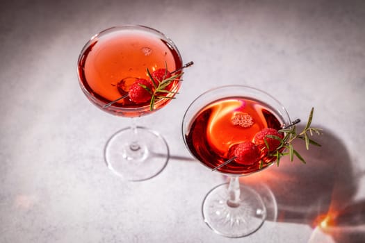 Two refreshing cocktails garnished with fresh raspberries and rosemary sprigs, served in elegant glasses on a grey table, creating a stylish and inviting scene