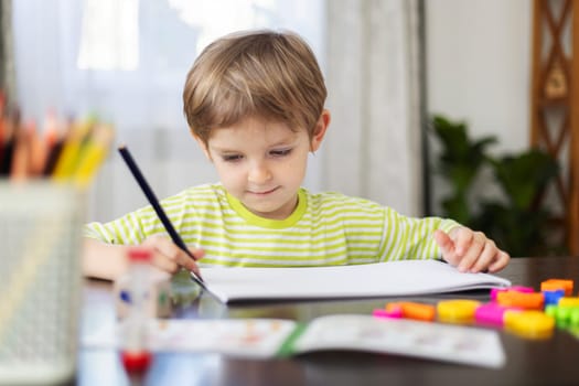 A young boy in a striped green and white T-shirt sits at a wooden table, drawing with a pencil on a white sheet of paper. Colored pencils, markers, and art supplies are scattered around him in a bright indoor setting
