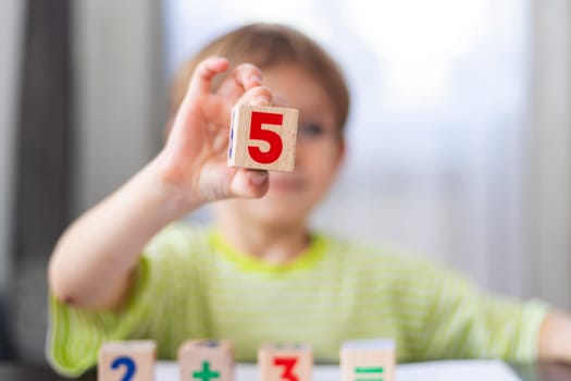 Child wearing a green striped shirt holding a wooden block with the number five printed on it. Other numbered blocks are on the table. Blurred background suggests a learning environment.