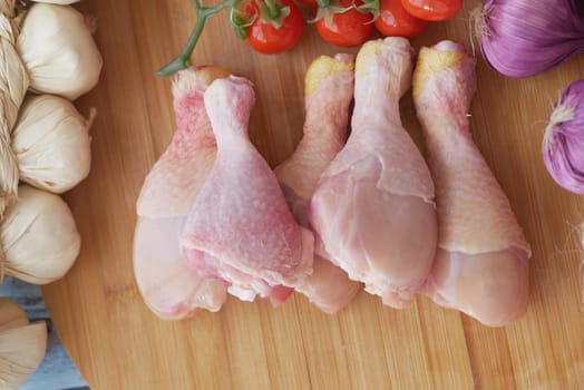 Juicy chicken drumsticks beside fresh tomatoes and garlic on wood.