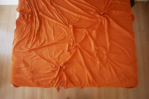 An unmade bed features a bright orange bedspread on a wooden floor.