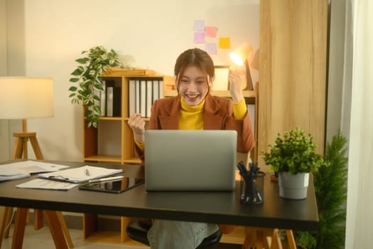 Excited young businesswoman looking at her laptop and celebrating success at work.