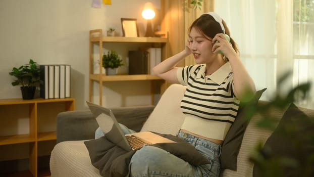 Young asian woman wearing wireless headphones and working on her laptop in living room.