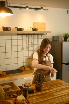Smiling young woman grinding coffee beans with hand grinder in a kitchen.