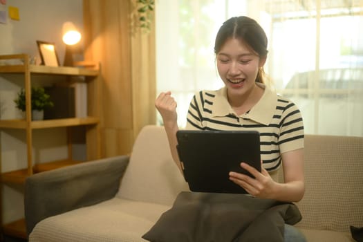 Cheerful young woman sitting on couch raising her fist in excitement while looking at her digital tablet.