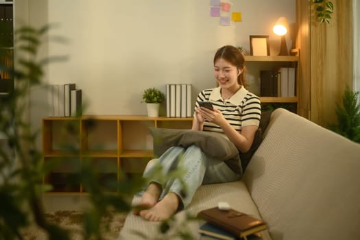 Smiling young asian woman enjoying social media on smartphone sitting on couch at home.