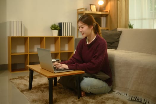 Smiling young woman in a maroon sweater using laptop in a cozy living room.