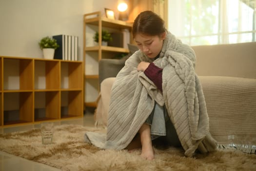 Sick woman sitting on the floor wrapped in a gray blanket. Illness and home recovery concept.