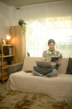 Relaxed young asian woman in casual clothes reading book on couch at home.