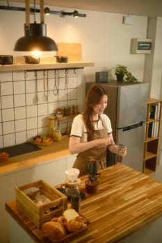 Smiling young woman grinding coffee beans with hand grinder in a kitchen.