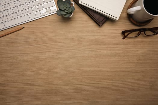 Top view shot of wooden desk with wireless keyboard, notebook, a cup of coffee and eyeglasses.