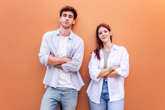 happy young couple of man and woman standing in front of orange wall looking at camera with arms crossed, concept of relationship and friendship