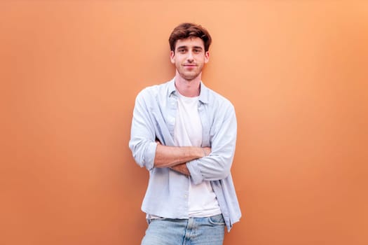 smiling young man standing in front of orange wall looking at camera with his arms crossed, he is wearing a white shirt and blue jeans