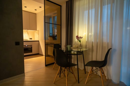 Modern apartment's dimly lit dining area. Glass-topped table with four black chairs sits near a glass-paned doorway leading to a kitchen. Sheer curtains frame a window.