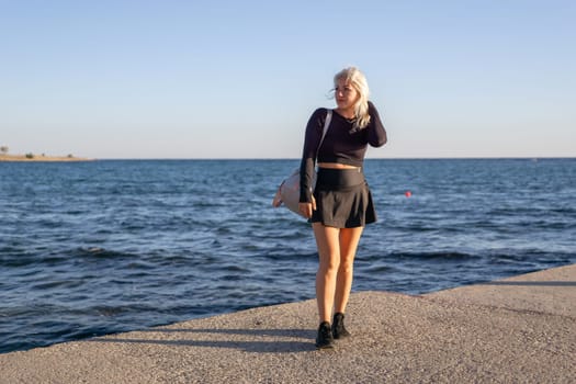 A blonde woman in a black skirt and top stands by the ocean, looking at the camera with a smile. She is holding a bag in her right hand.