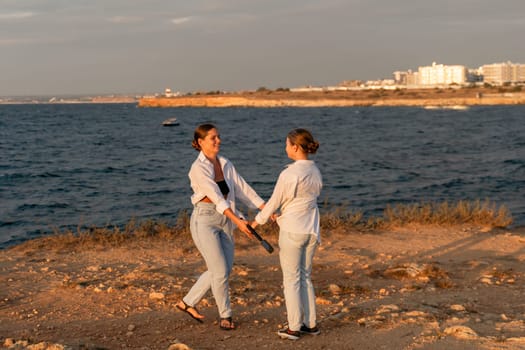 Two women in white shirts and blue jeans stand on a rocky beach, holding hands and smiling, looking at each other, with a calm blue sea and a distant city skyline in the background.
