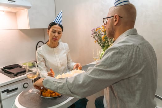 Here's a caption for the image: A man and woman celebrate a birthday indoors. They are seated at a small table with a birthday cake and party hats. A bouquet of flowers sits nearby.