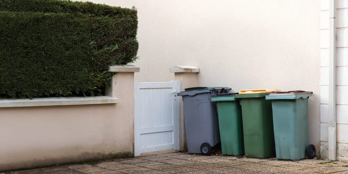 Different colored waste bins are neatly arranged along the side of a residential street. These containers include black, green, blue, and yellow bins for recycling and waste disposal.