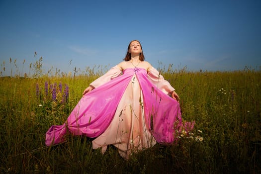 A woman stands in a vibrant field, wearing a flowing pink dress, with arms outstretched, embracing the tranquility of nature under blue skies.