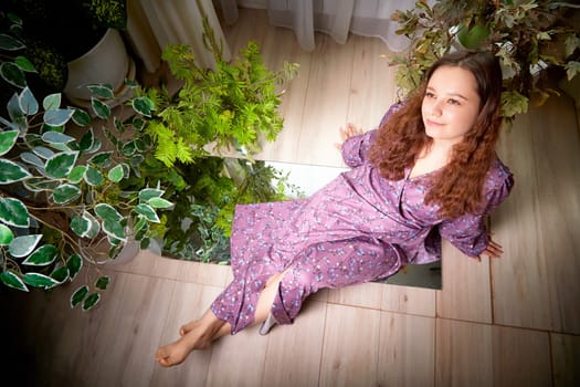 A young woman with curly hair sits calmly amidst various indoor plants, showcasing her purple dress in a beautifully lit setting.
